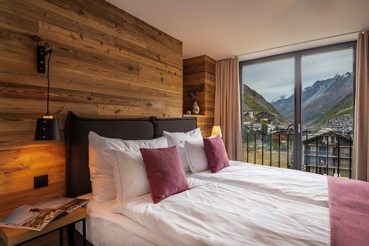Ein Schlafzimmer mit einem hölzernen Kopfteil, einem großen Bett mit weißen und rosafarbenen Kissen, einem Nachttisch mit Lampe und Bergblick durch ein großes Fenster.