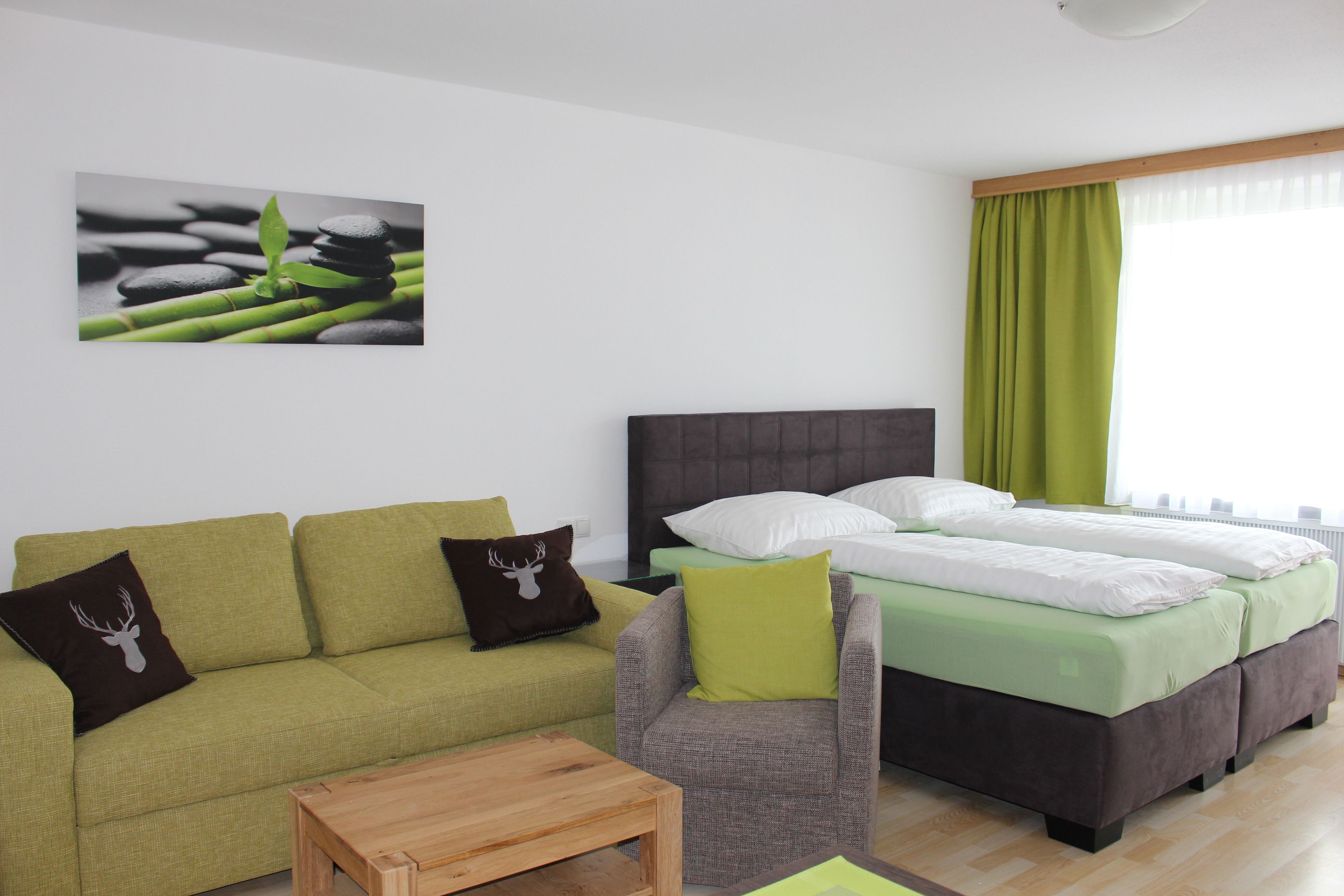 A modern hotel room with a green sofa, a grey armchair, a wooden coffee table, and a bed with green bedding.