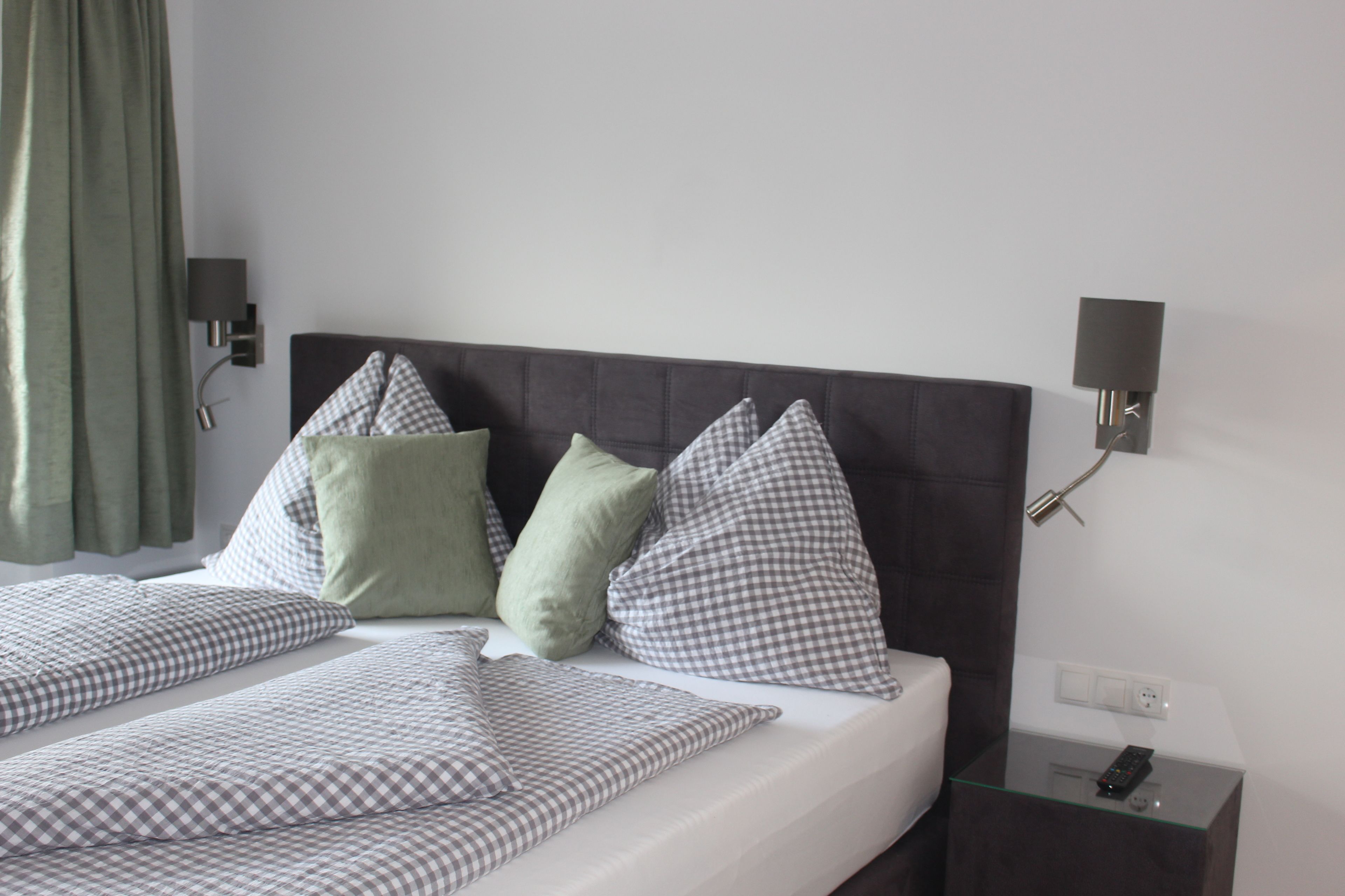 A neatly made bed with checkered and solid pillows, a glass side table with a remote control, and a wall-mounted lamp.