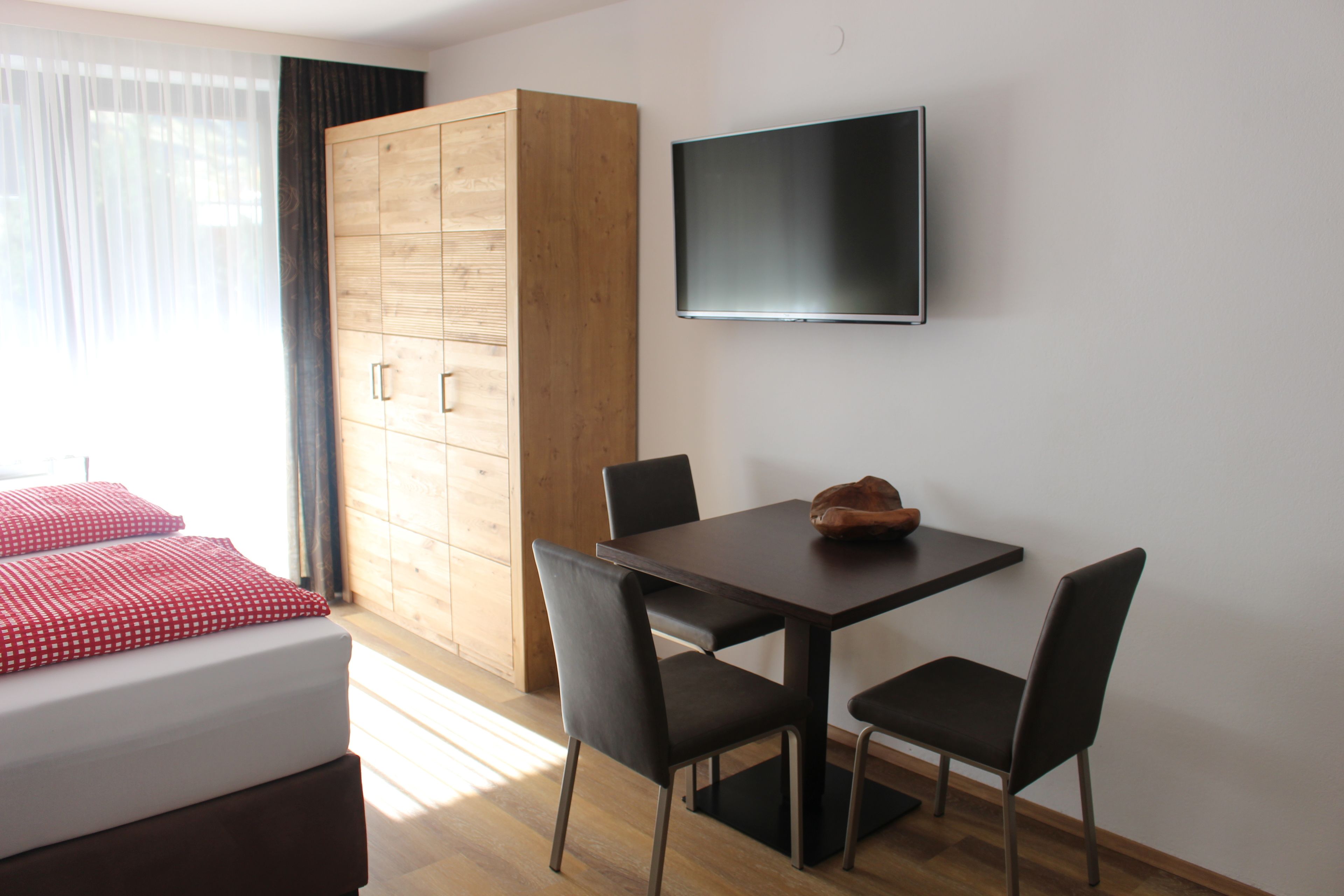 Een kamer met een bed, een houten garderobe, een tafel met stoelen en een wandtelevisie.