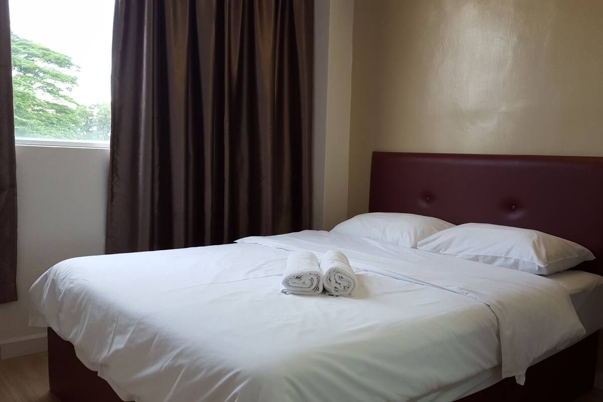 Un lit bien fait, avec du linge de lit blanc et deux serviettes enroulées, une tête de lit en bois et une table de chevet avec un tiroir.