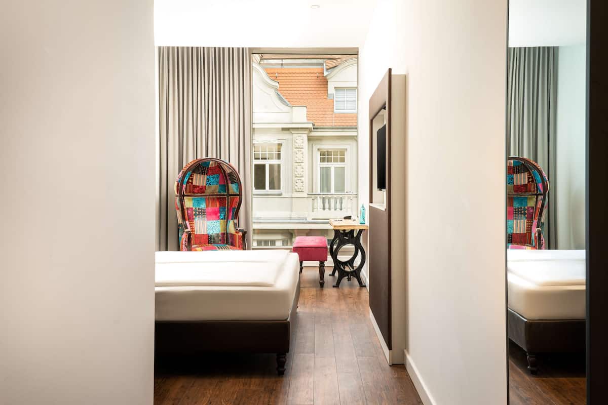 A narrow hotel room with two beds, a small table, and a view of a building through a window.