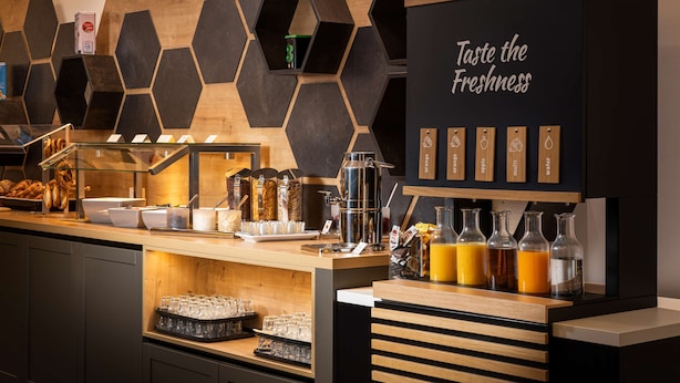 A breakfast bar with hexagonal wall decor, a sign that says "Taste the Freshness," and various food and drink items. - elaya hotel leipzig city center (Leipzig)