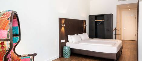 Un dormitorio con una cama, una silla de madera con tapicería colorida, un cabecero oscuro y un pequeño jarrón decorativo.