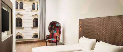 A hotel room with a large bed, a colorful armchair, a television, and a view of a building with arched windows.