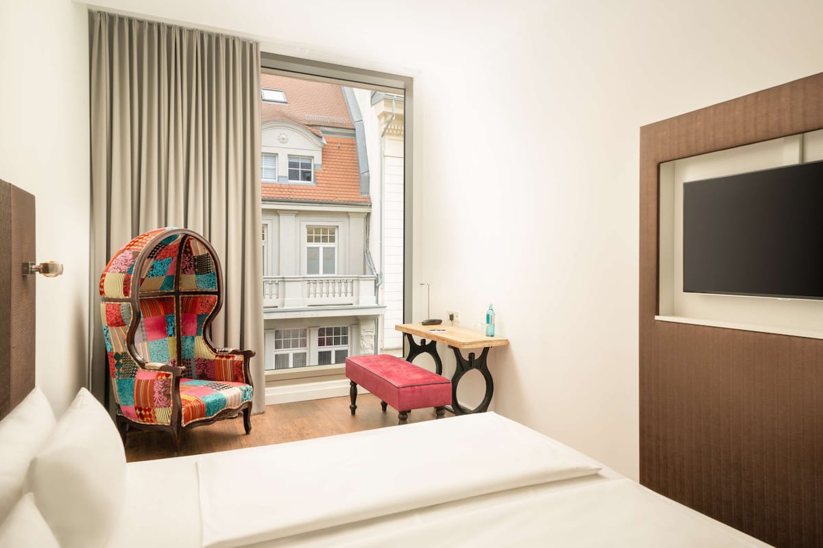 A hotel room with a large window, a colorful armchair, a small table, a red ottoman, and a bed.
