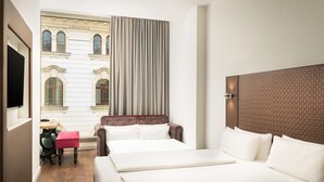 A hotel room with a large bed, a small table with a chair, and a view of a historic building through a window. - elaya hotel leipzig city center (Leipzig)