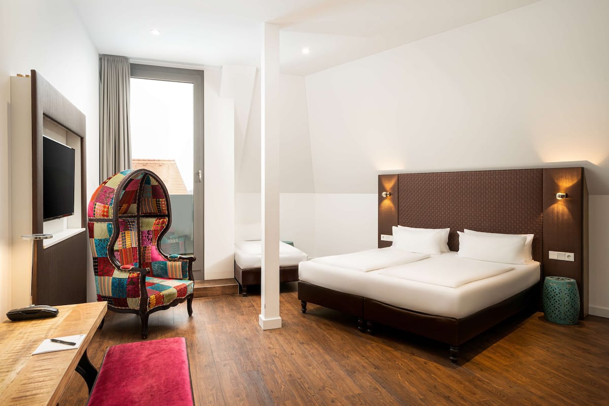 A modern hotel room with a large bed, a colorful armchair, a desk with a chair, and a television.