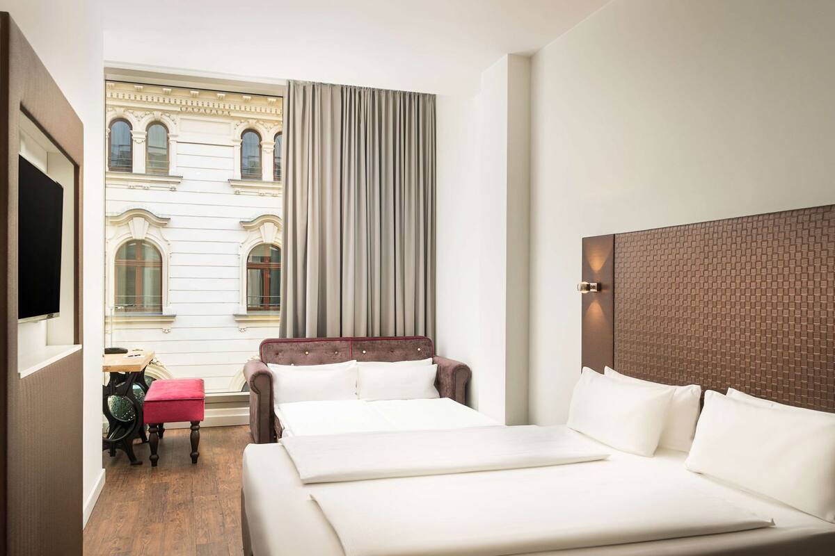 A hotel room with a large bed, a small table, a chair, and a view of a building with arched windows.