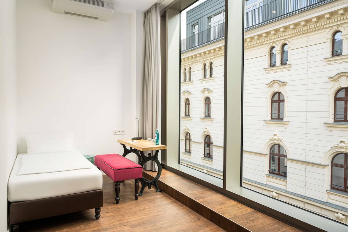 A hotel room with a bed, a red bench, a small table, and a view of a historic building through large windows.