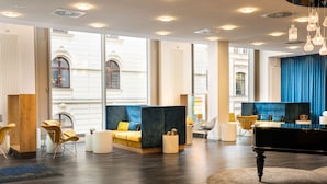 A modern hotel lobby with a grand piano, comfortable seating areas, and large windows offering views of the building's architecture. - elaya hotel leipzig city center (Leipzig)