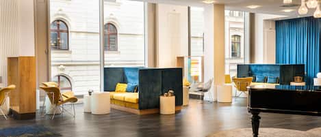 A modern hotel lobby with a grand piano, comfortable seating areas, and large windows offering views of the building's architecture.