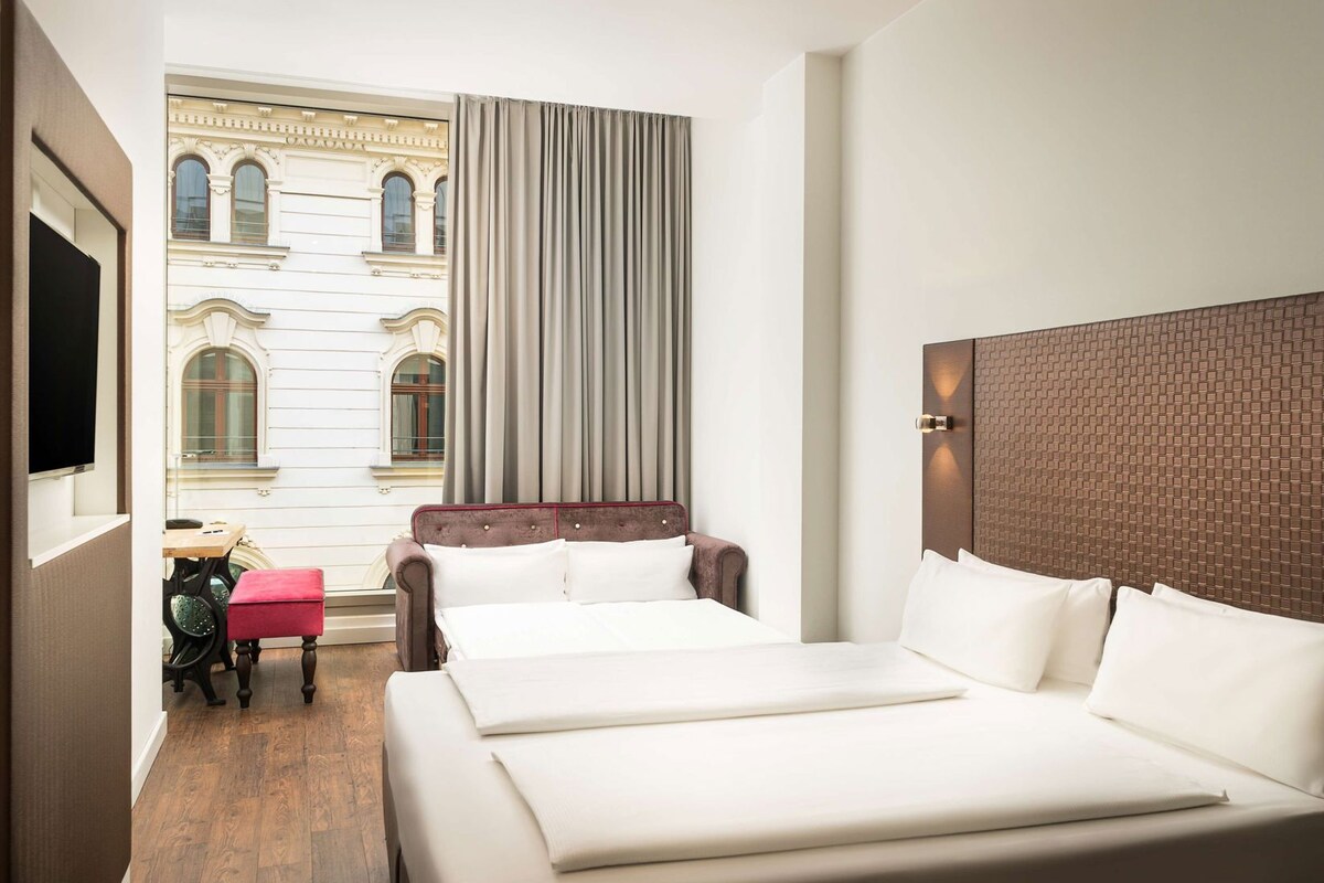 A hotel room with a large bed, a small table, a chair, and a view of a building with arched windows.