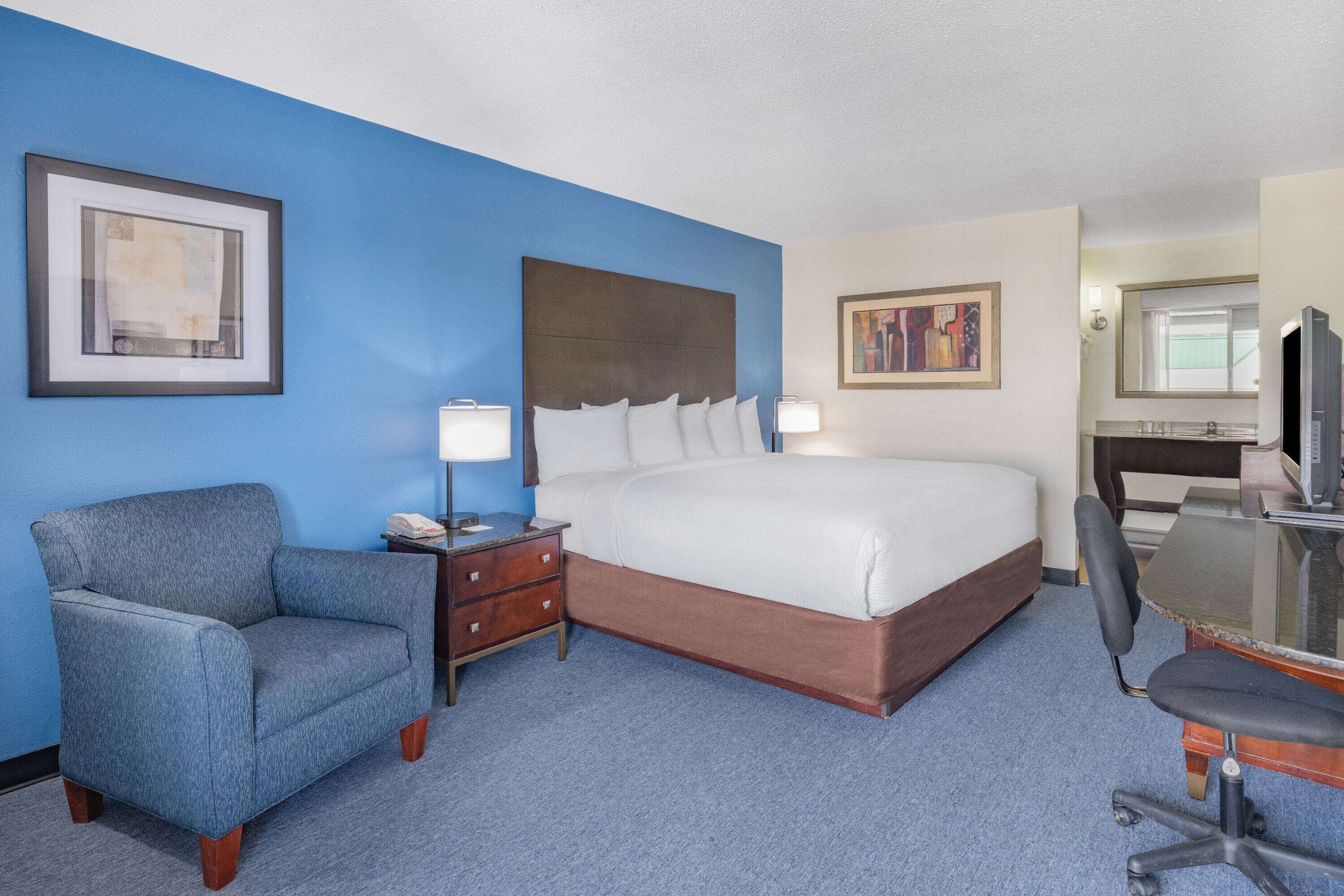 A hotel room with a bed, a desk with a chair, a small table with a lamp, a chair, and two framed pictures on the walls.