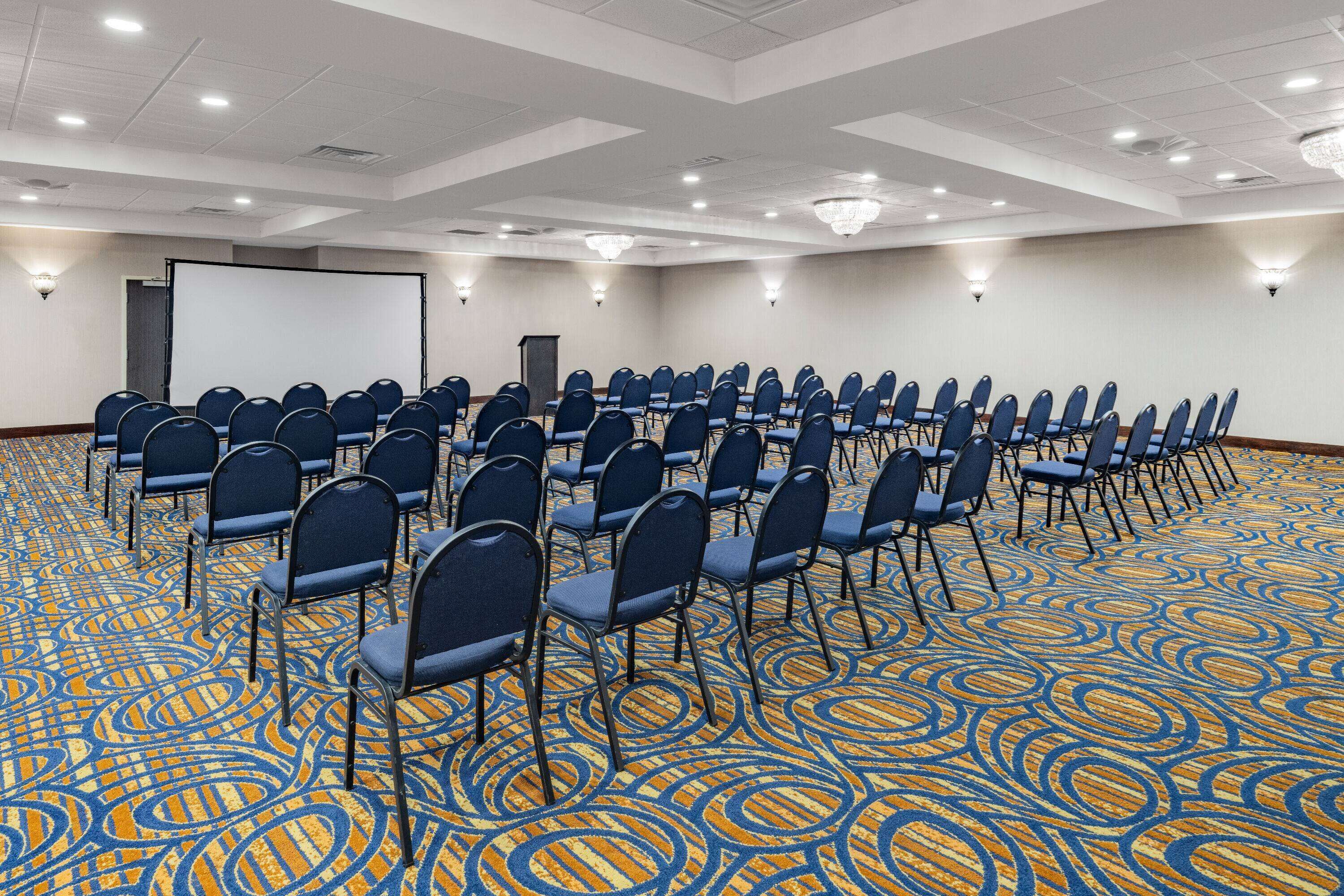 Une salle de conférence avec des rangées de chaises, un écran de projection et un sol recouvert de moquette.
