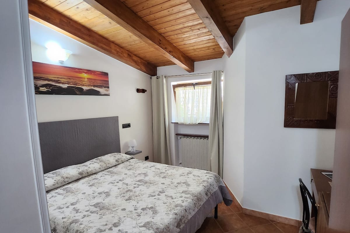 Une chambre avec un plafond en bois, un lit recouvert d’une literie à motifs floraux, une fenêtre avec des rideaux, un radiateur et une petite table avec une chaise.