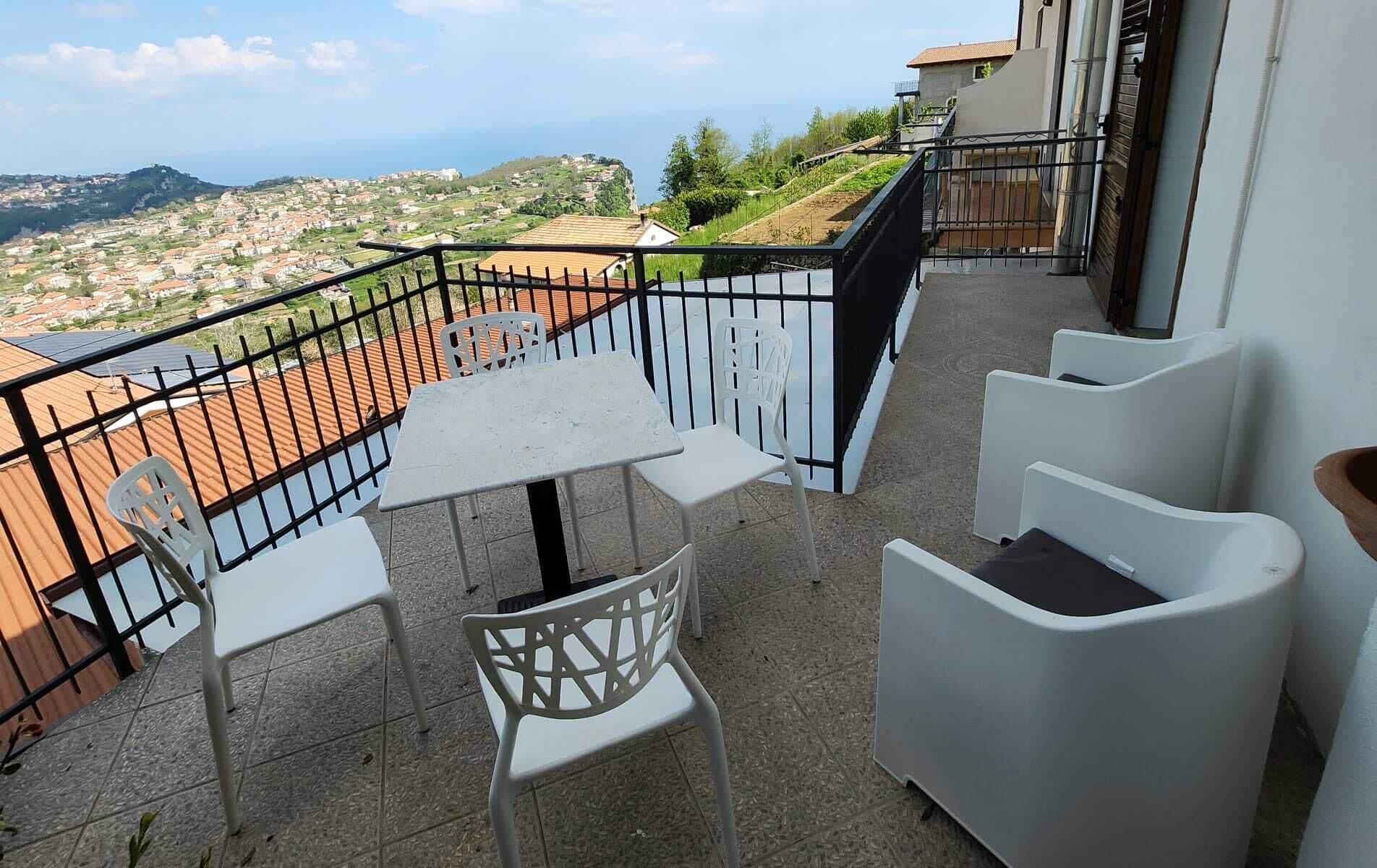 A balcony with a table and chairs, a view of a hillside town, and a black metal railing.