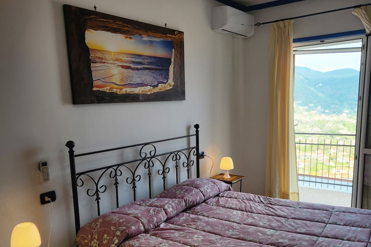 Une chambre à coucher avec un lit, une table de chevet avec une lampe, un tableau au mur et une fenêtre donnant sur l’extérieur.