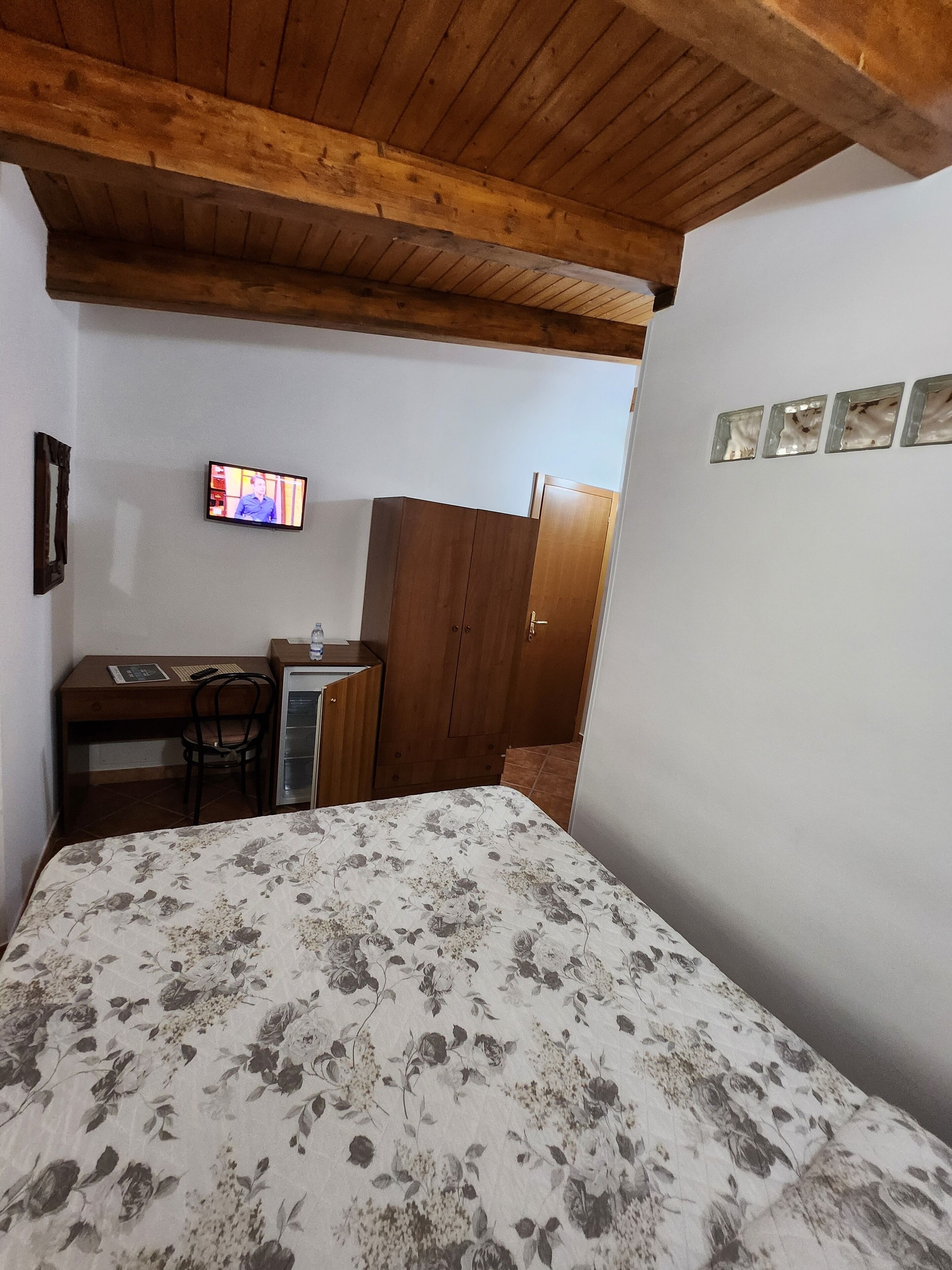 A bedroom with a wooden ceiling, a bed with a floral bedspread, a desk with a chair, a television, and a wardrobe.