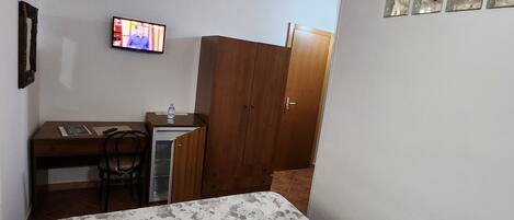 A bedroom with a wooden ceiling, a bed with a floral bedspread, a desk with a chair, a television, and a wardrobe.