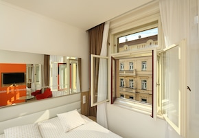 A hotel room with a bed, a TV, a window with curtains, and a view of a building.