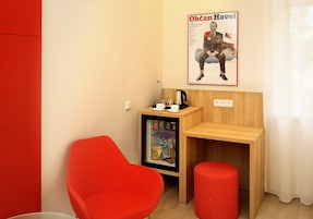 Quarto pequeno com uma cadeira vermelha, escrivaninha de madeira e um refrigerador com lanches e bebidas.