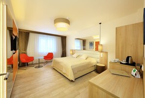 Quarto de hotel moderno com uma cama grande, uma escrivaninha com um laptop, uma máquina de café e uma televisão. Há duas cadeiras vermelhas e uma mesa pequena.