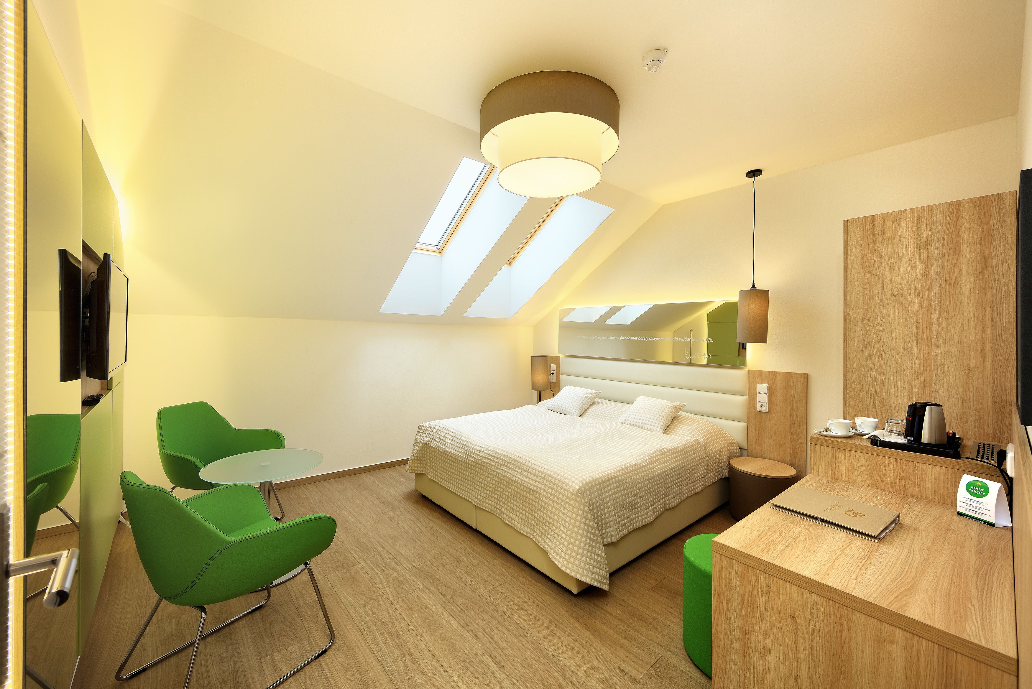 A modern hotel room with a bed, green chairs, a desk, a mirror, and skylights.