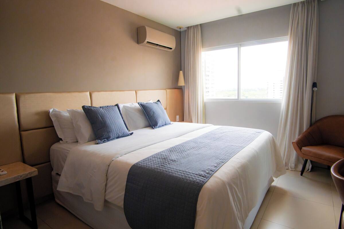 Een hotelkamer met een groot bed, nachtkastje, stoel en raam met gordijnen.