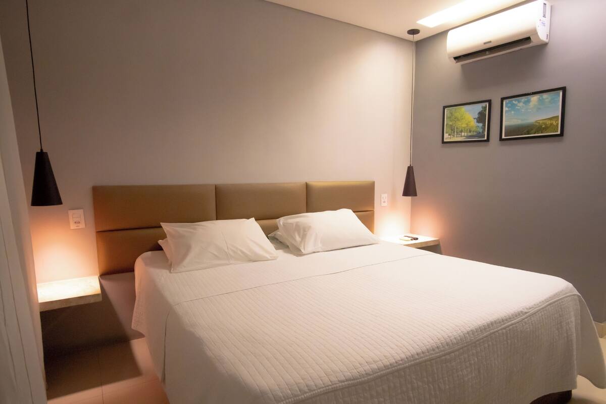 Een netjes opgemaakt bed met hoofdeinde, twee nachtkastjes met lampen, en twee ingelijste foto's aan de muur.