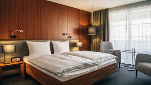 Habitación de hotel con cama de madera, dos lámparas de noche, una silla, una mesita y un gran ventanal con cortinas.