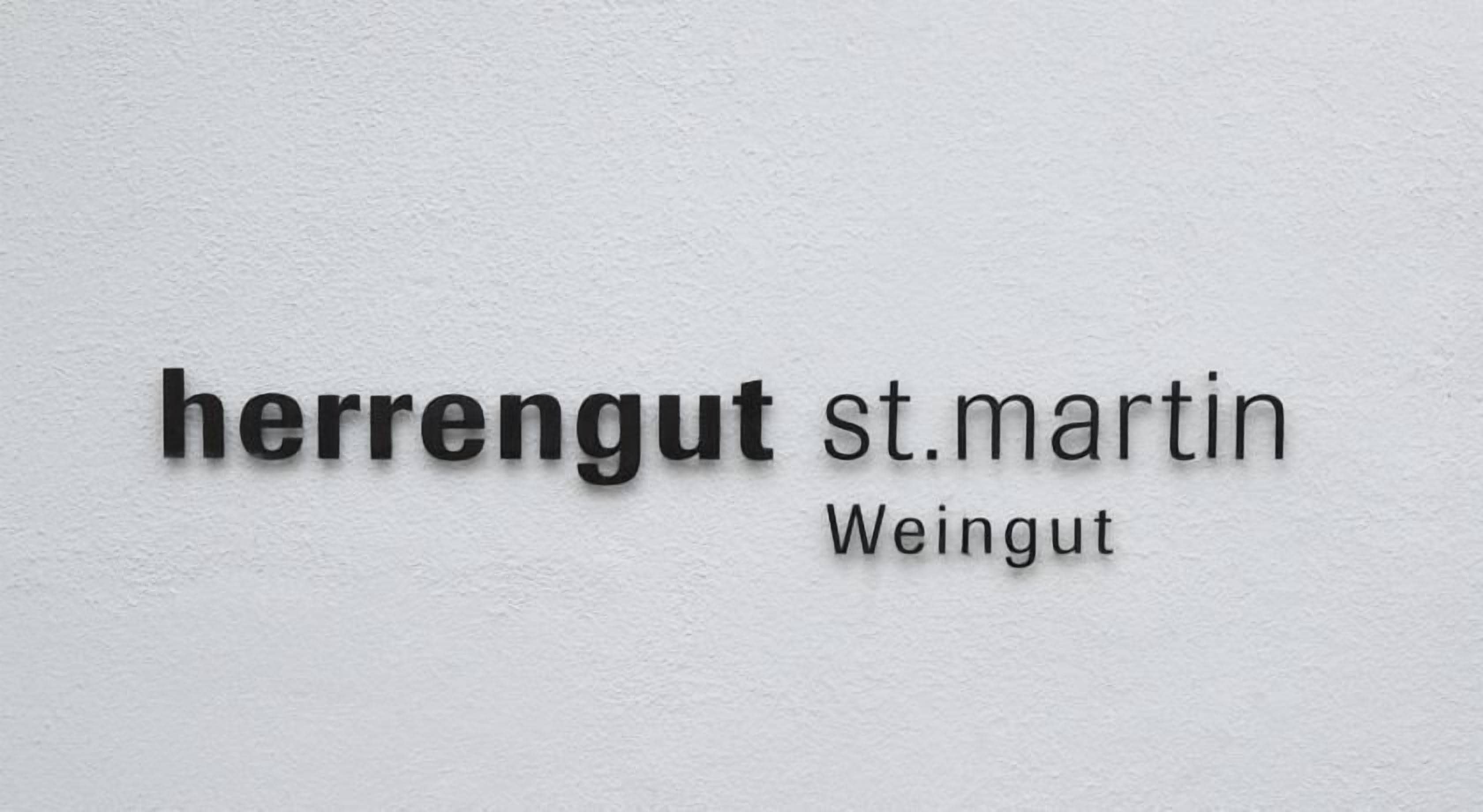 Ein Schild mit dem Text 'herrengut st.martin' und 'Weingut' vor weißem Hintergrund.