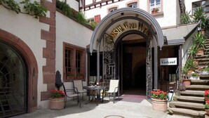 Un edificio tradicional europeo con fachada de entramado de madera, una entrada arqueada, asientos exteriores y plantas en macetas.