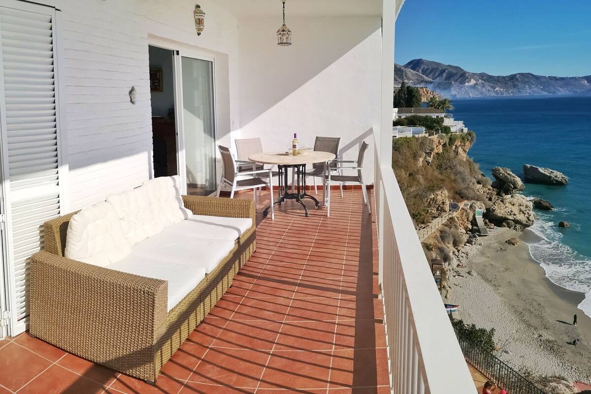 Un balcón con un sofá de mimbre, una mesa con sillas y vistas al mar y a las montañas.