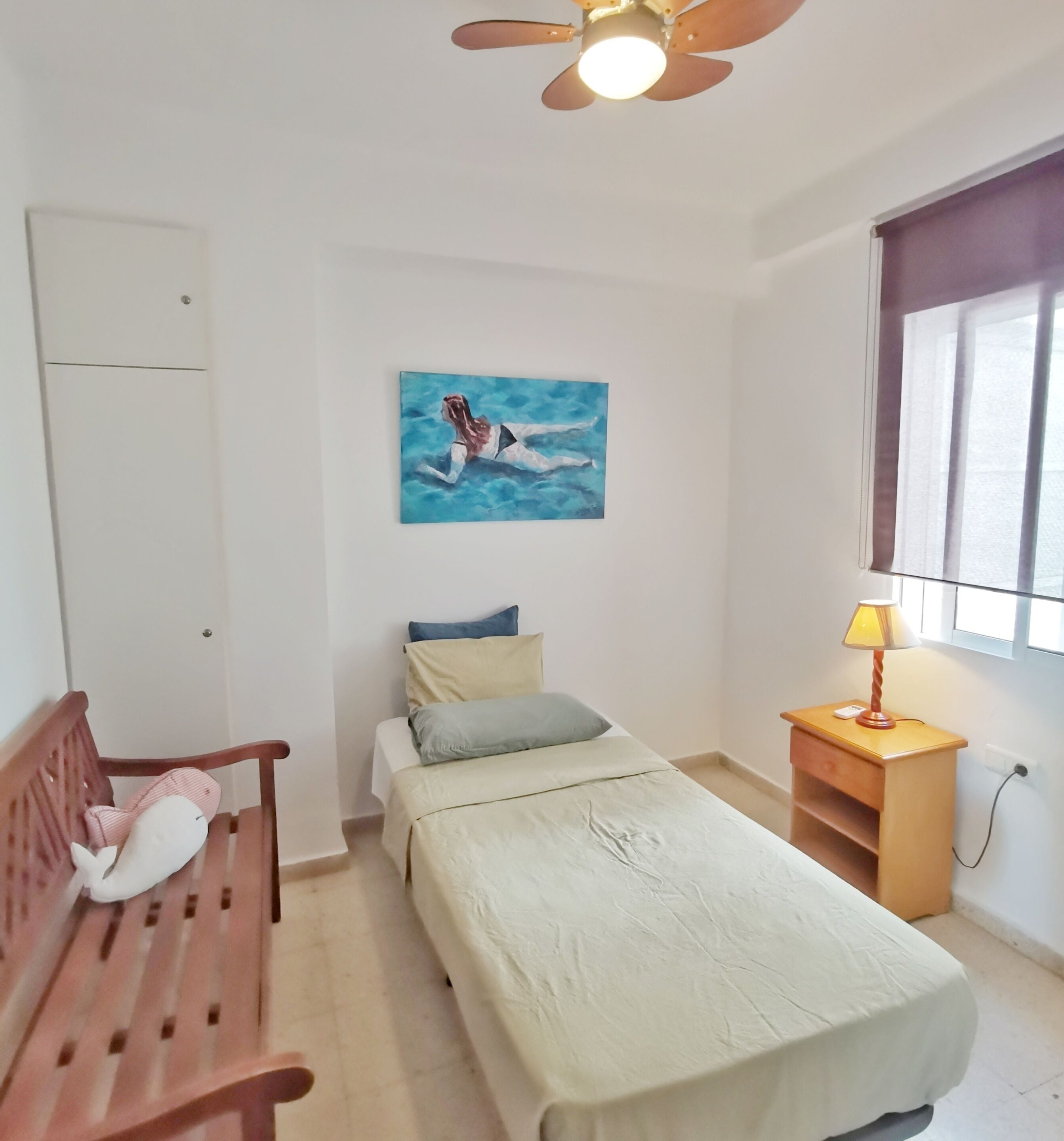 Una camera da letto con un letto, una sedia di legno, un comodino con una lampada, un quadro appeso e una finestra con tende.