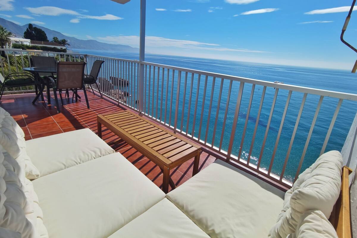 Un balcón con mesa de madera y asientos blancos acolchados, con vista al mar azul cristalino y montañas lejanas.
