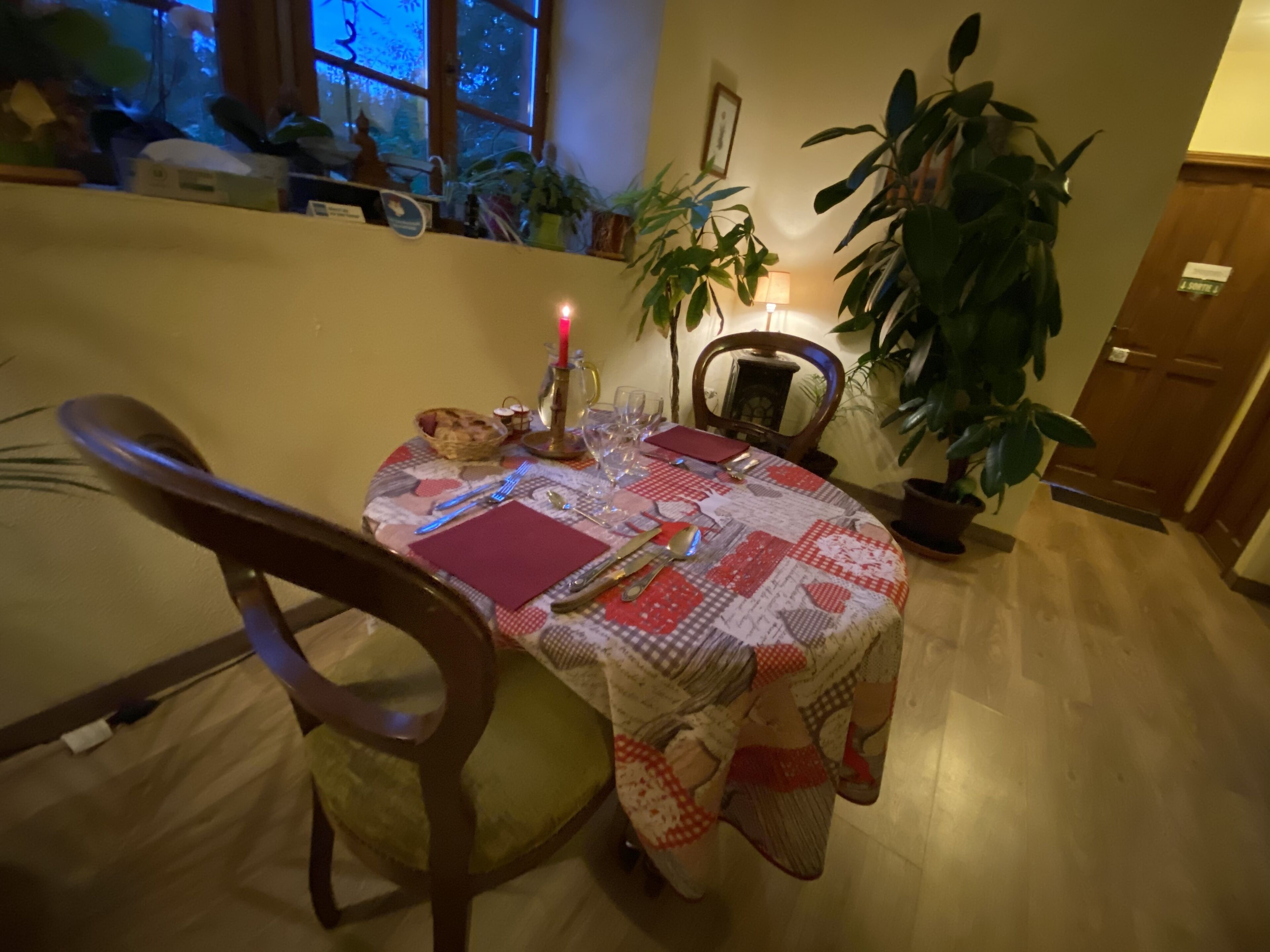Un comedor con una mesa redonda dispuesta para dos personas, una vela y varios utensilios de mesa. Hay sillas, una planta en maceta y una ventana al fondo.