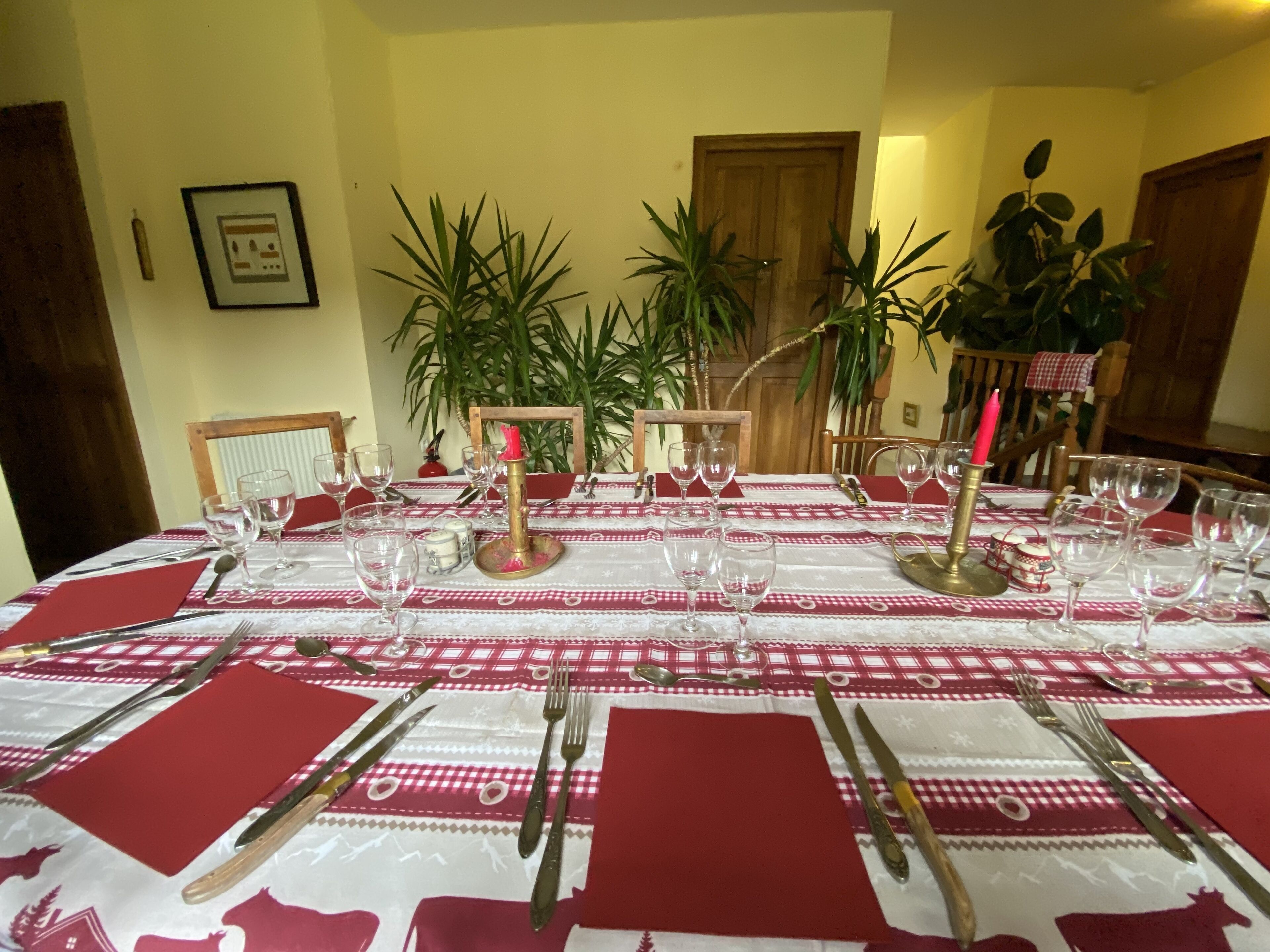 Mesa de comedor dispuesta con mantel rojo y blanco, rodeada de sillas, con plantas en macetas y una puerta de madera al fondo.