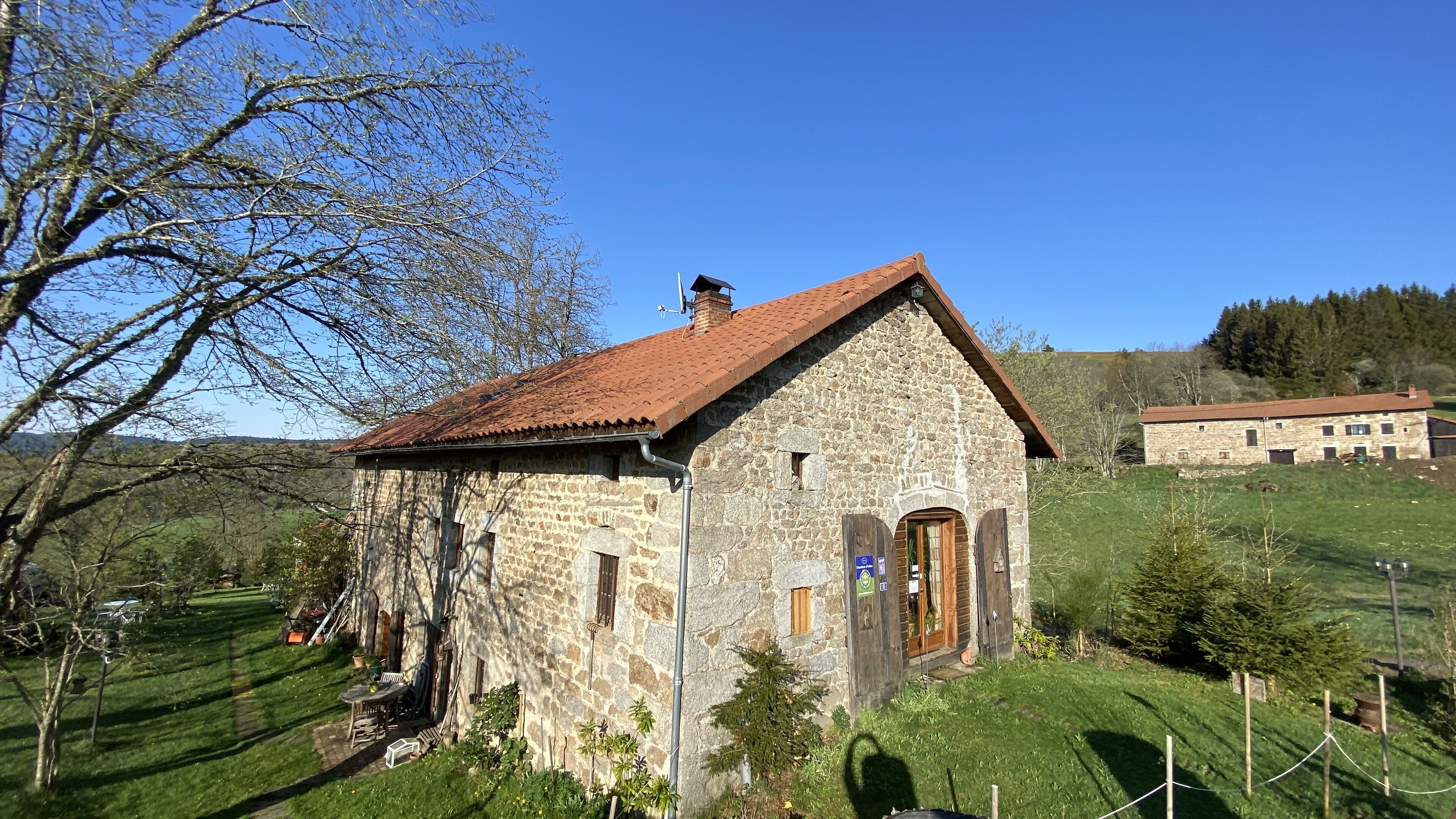 Un bâtiment en pierre avec un toit recouvert de tuiles rouges, une porte en bois et un petit jardin agrémenté d’une table et de chaises.