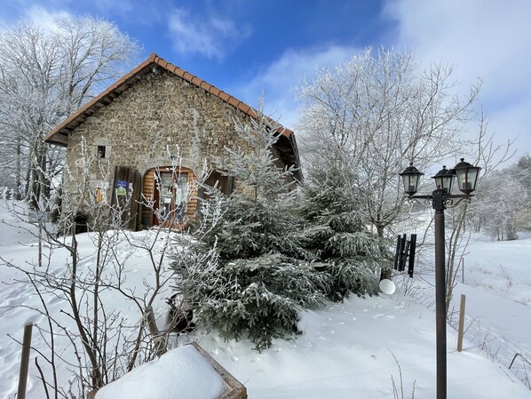 A stone building with a wooden door and windows, surrounded by snow-covered trees and a snow-covered ground.