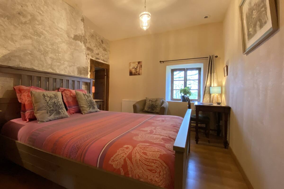 Ein Schlafzimmer mit einem hölzernen Bett, einem Nachttisch, einem Schreibtisch, einem Stuhl, einem Fenster mit Vorhängen und einer Pendelleuchte.