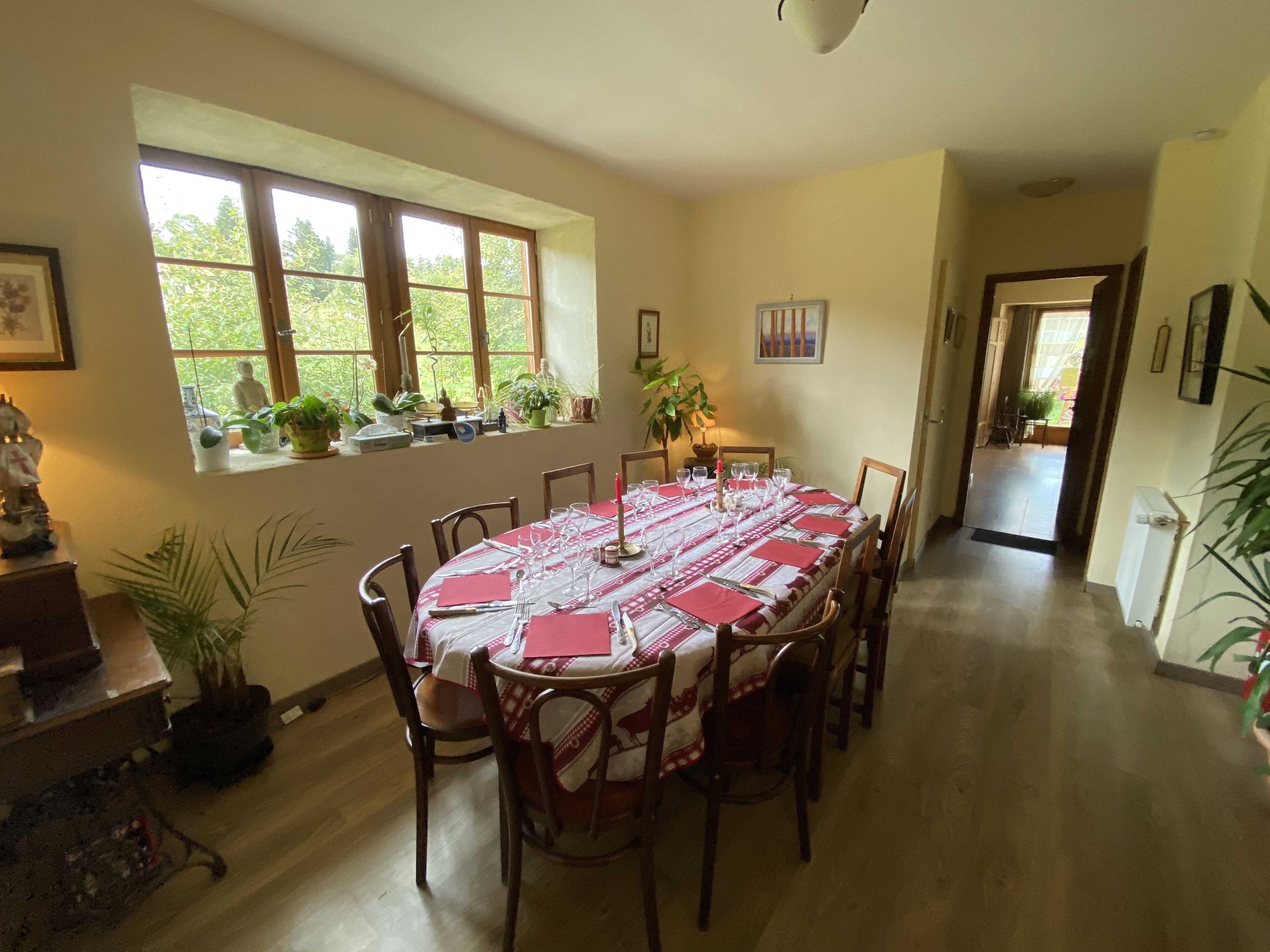 A dining room with a round table set for six, surrounded by wooden chairs. There are potted plants on the windowsill and a small table with decorative items.