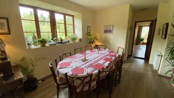 A dining room with a round table set for six, surrounded by wooden chairs. There are potted plants on the windowsill and a small table with decorative items.