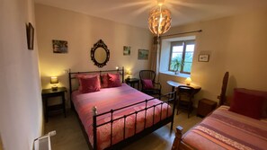 A bedroom with a bed, bedside tables, a chair, a desk, a window, and a chandelier. - Jasserie Les Airelles (Saint-Antheme)