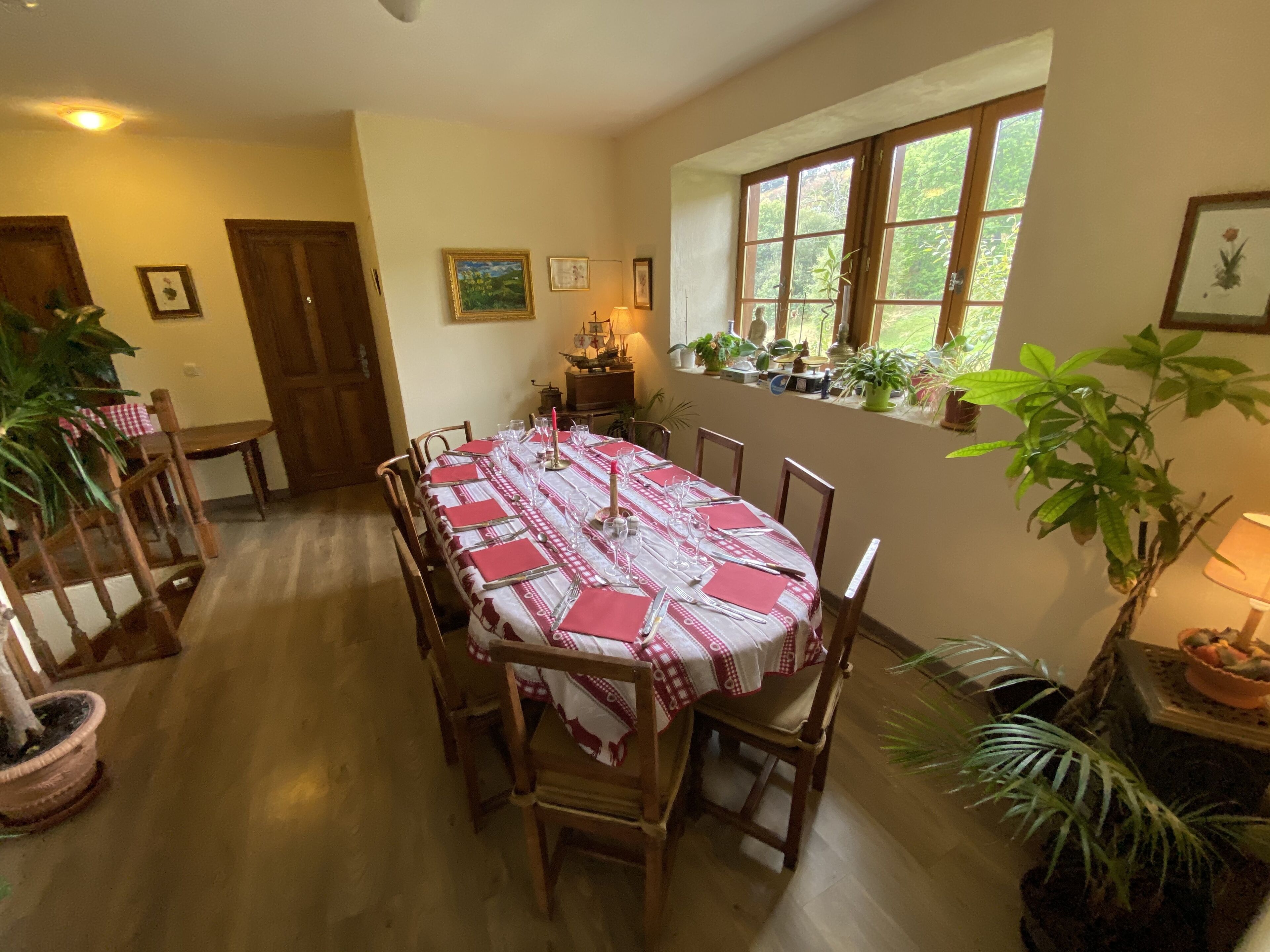 A dining room with a round table set for six, surrounded by wooden chairs. There are potted plants on the windowsill and a door to the left.