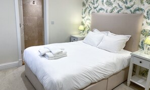 A neatly made bed with white linens and towels, a nightstand with a lamp, and a shower area with a towel rack.