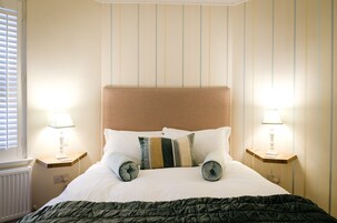 Una cama bien hecha con cabecera de madera, dos mesitas de noche con lámparas y una ventana con persianas.