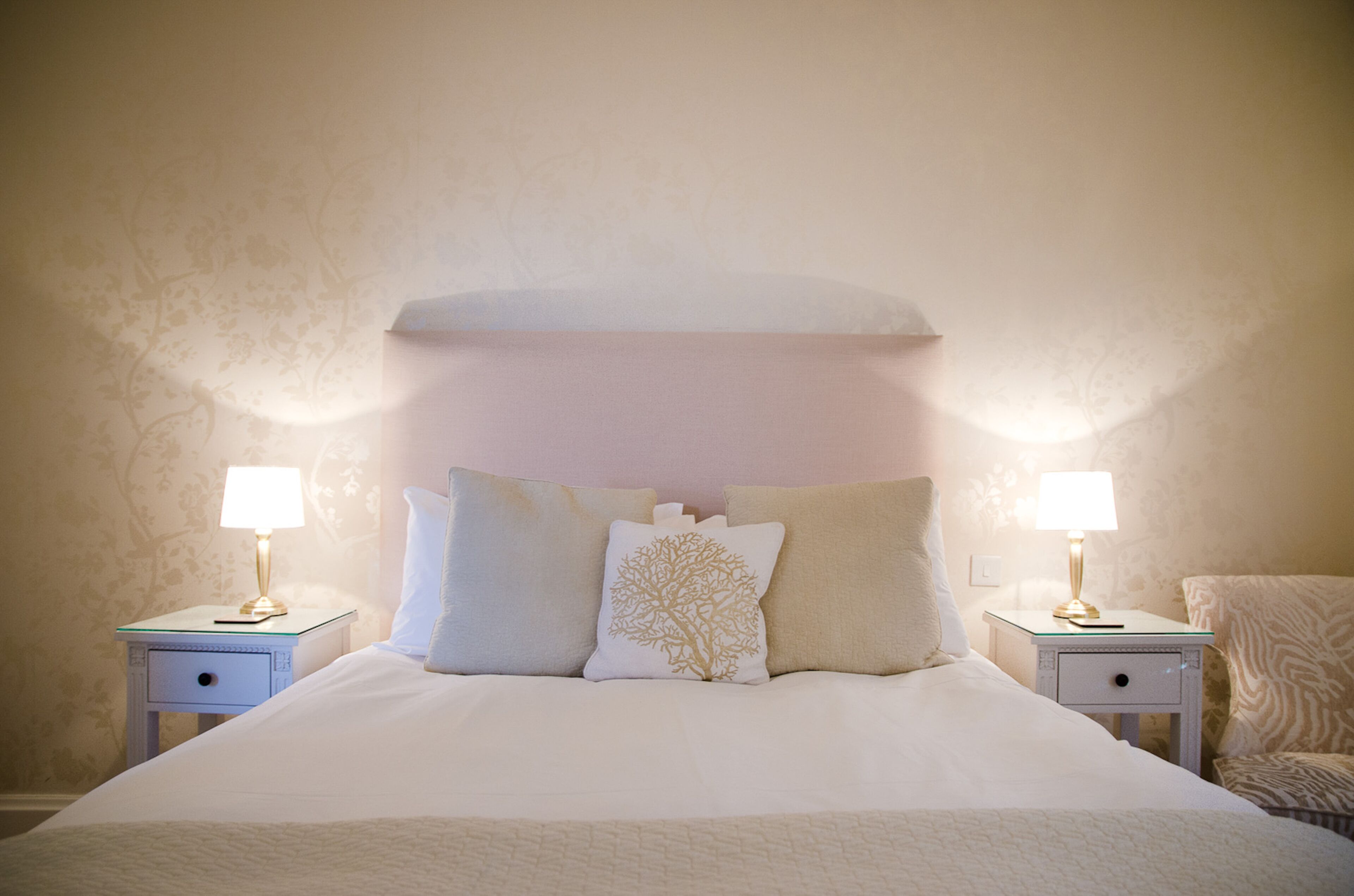 A neatly made bed with white bedding and a tree-patterned pillow, flanked by two bedside tables with lamps, in a room with patterned wallpaper.