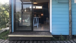 Un edificio pequeño de una sola planta con puerta de vidrio corredera, terraza de madera y un interior sencillo con una mesa y sillas.
