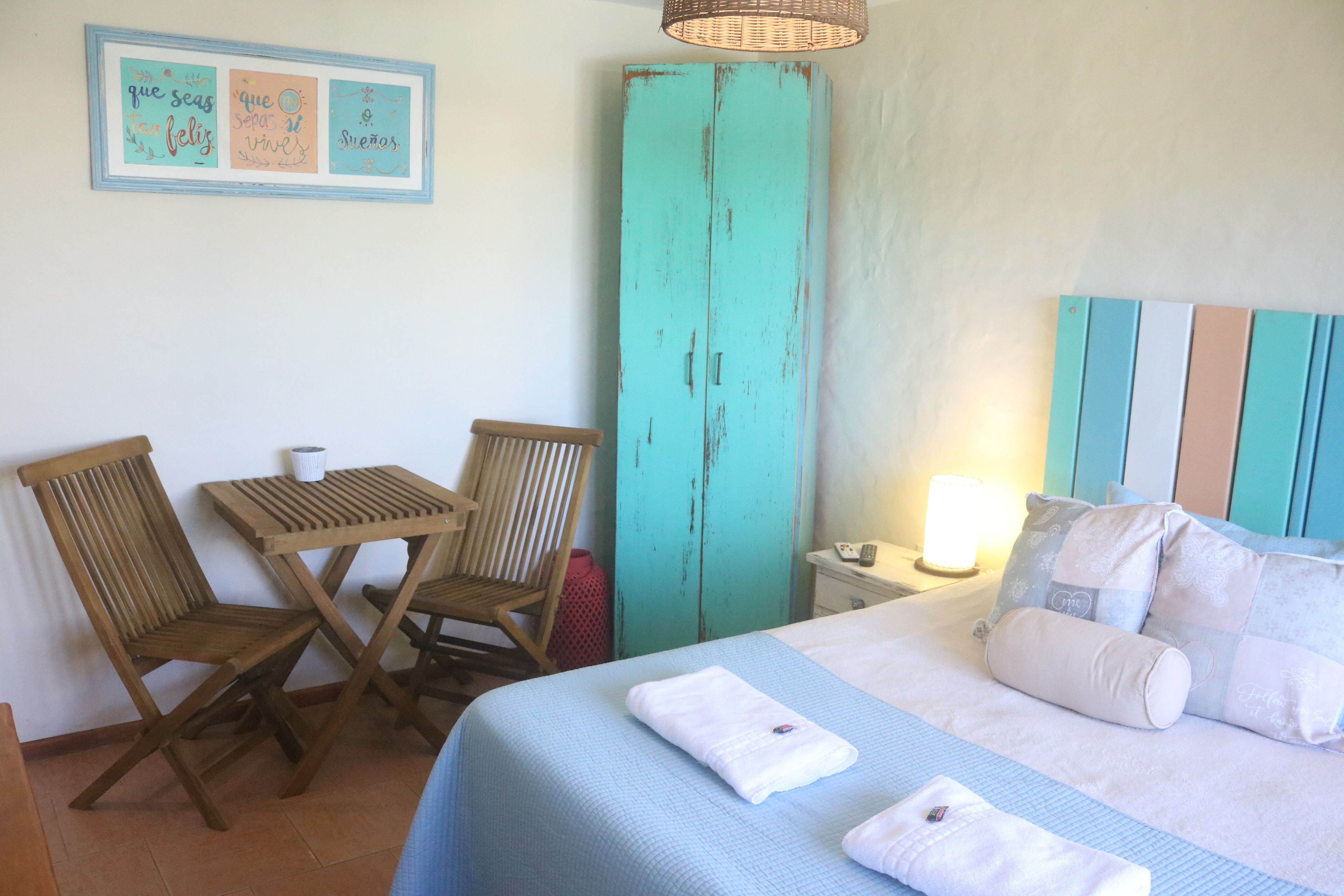 A room with a bed, a wooden table with chairs, a teal wardrobe, and a wall with framed text.